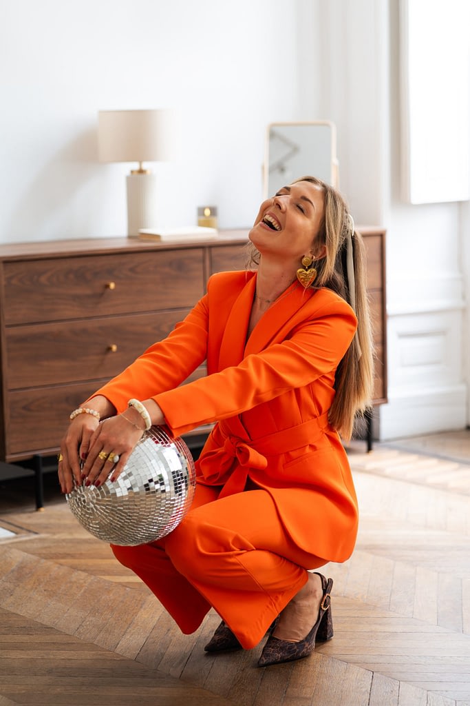 photo de Meije en tailleur orange riant les yeux fermés, accroupie avec une boule à facettes entre les mains dans un intérieur lumineux