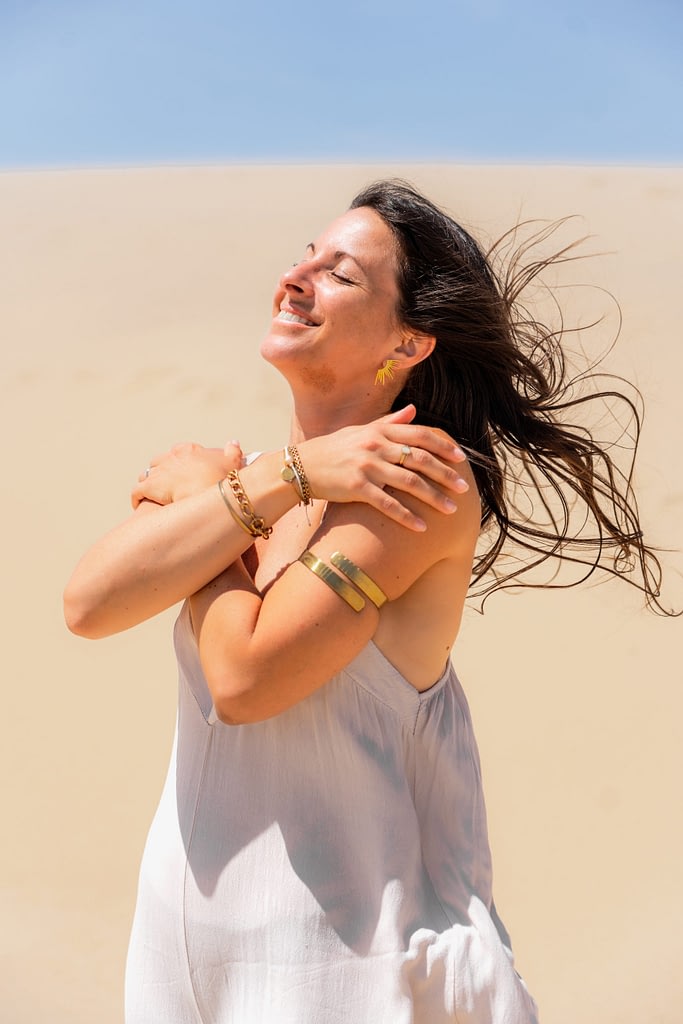 Femme souriante les yeux fermés, les bras croisés sur ses épaules et les cheveux au vent, devant une dune ensoleillée.
